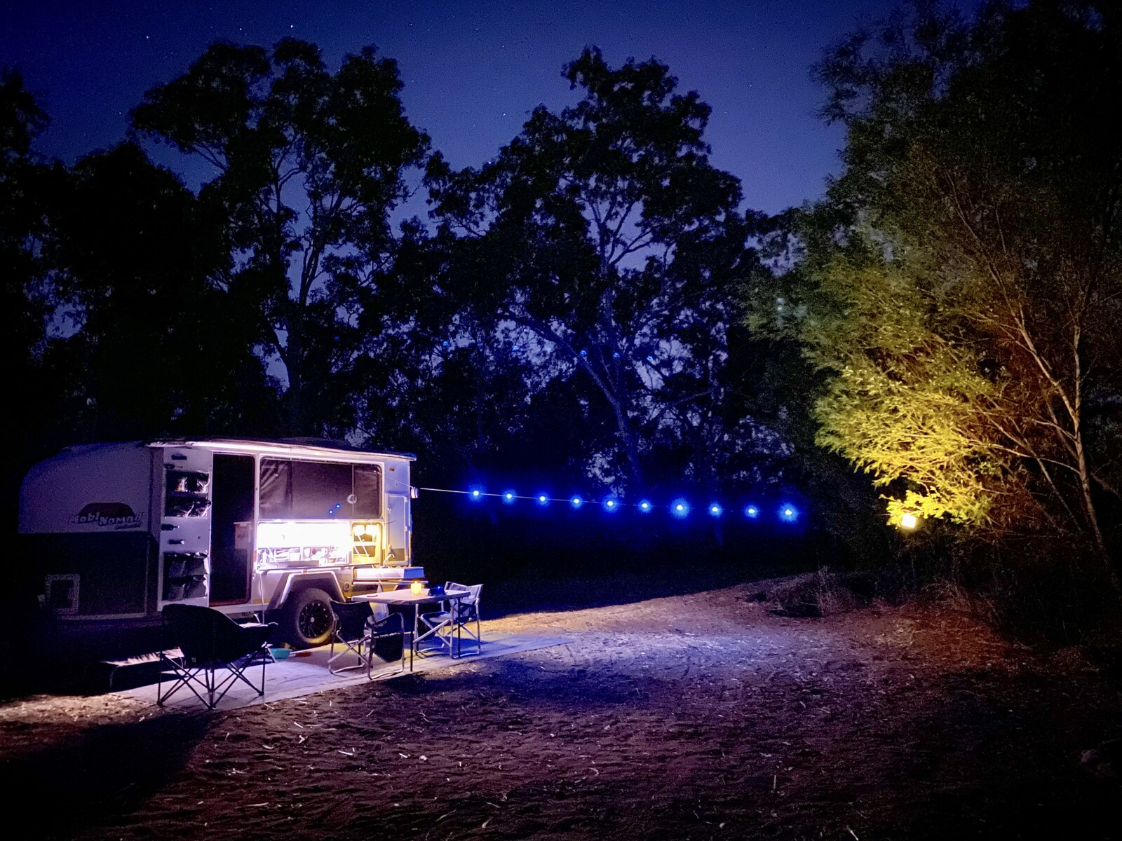 A Mobi-Nomad camper trailer set up for dinner under the stars with fairy lights strung from a tree